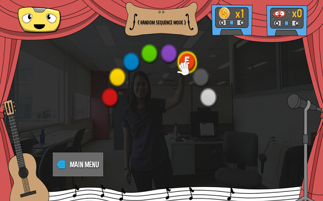 A cartoonish video game screenshot depicts a rainbow of circles on the centre of the screen. The circles are overlaid on a real picture of a smiling player. 'Random Sequence Mode' is written at the top of the screen. A white hand-shaped cursor appears on top of the player's left hand, hovering over a circle labeled 'E'. A robot smiles at the top of the screen. The game score is one coin and zero robots collected. A blue arrow at the bottom of the screen is labeled 'Main Menu'. A guitar and a microphone flank either side of the screen, which is framed with red curtains.